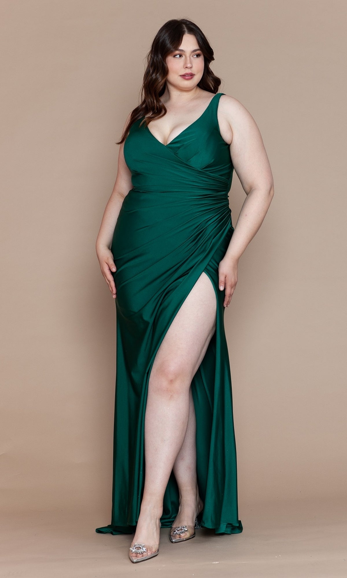 A woman models the Faux-Wrap Plus-Size Long Prom Dress W1114, featuring a sleeveless deep green design with a thigh-high slit and ruched waist, standing against a beige background.